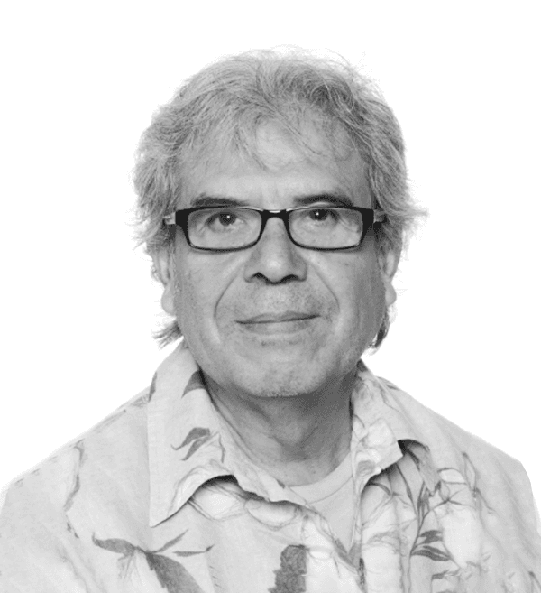 Portrait d'un homme âgé aux cheveux gris, portant des lunettes et une chemise à motifs sur fond blanc.