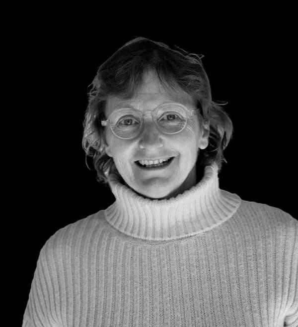 Lächelnde Frau mit schulterlangen Haaren, die eine runde Brille und einen dicken, gerippten weißen Rollkragenpullover trägt.