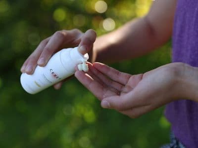Person drückt eine weiße Lotion aus einer Flasche mit der Aufschrift 'CERES' auf die offene Hand vor einem unscharfen grünen Hintergrund.