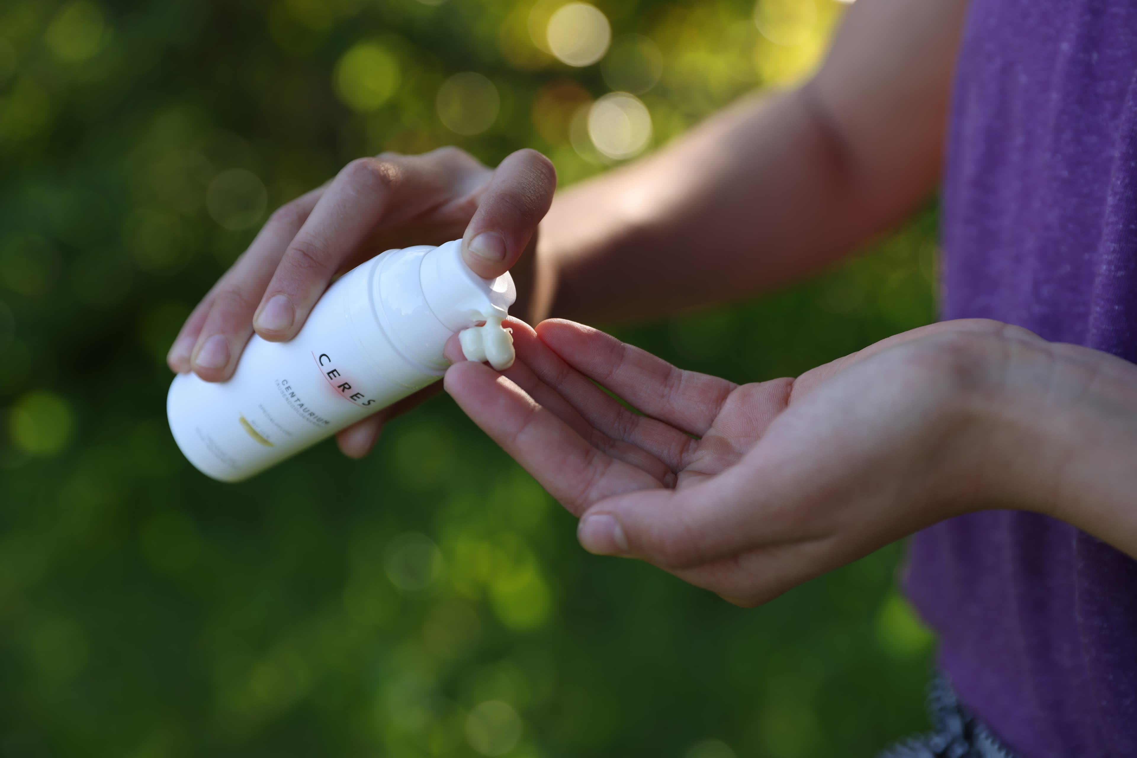Person drückt eine weiße Lotion aus einer Flasche mit der Aufschrift 'CERES' auf die offene Hand vor einem unscharfen grünen Hintergrund.